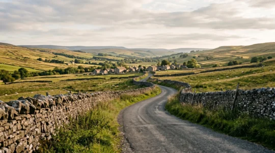 Winding single-track country road leading through traditional farming landscape with dry stone walls and distant village