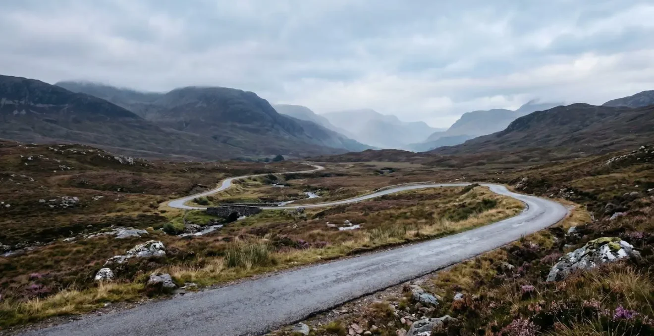 A dramatic winding single-track road cutting through misty Scottish Highlands with sweeping mountain vistas under moody atmospheric skies
