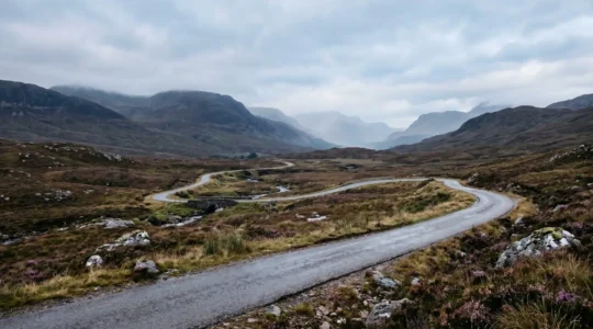 A dramatic winding single-track road cutting through misty Scottish Highlands with sweeping mountain vistas under moody atmospheric skies
